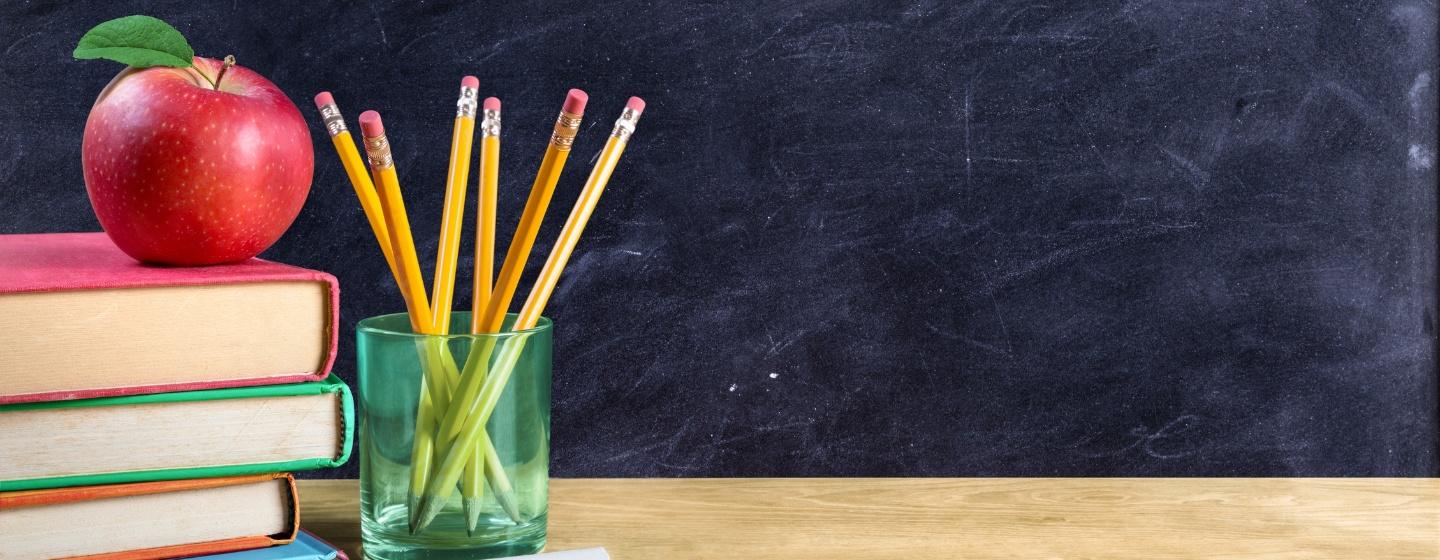 A stack of books with an apple on top is shown in front of a blackboard.